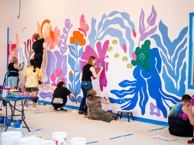 Painting of mural at Marie Equi Center - image shows four people standing, kneeling and sitting, with paintbrushes, painting a multicolored swirly mural on the wall