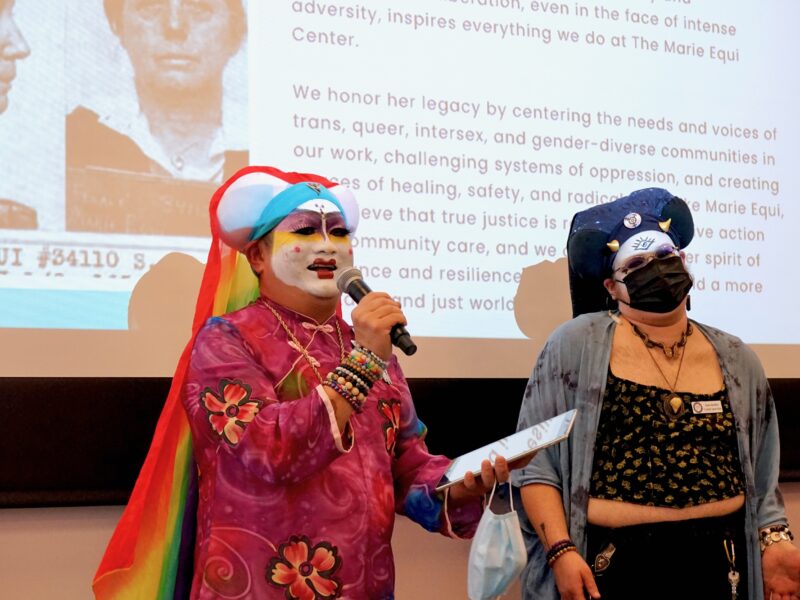 Two people wearing colorful makeup and clothing talk to an audience.