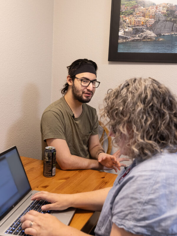 Jayson sits at a table with a person at a laptop.