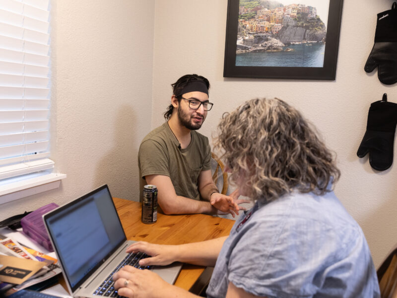 Jayson sits at a table with a person at a laptop.