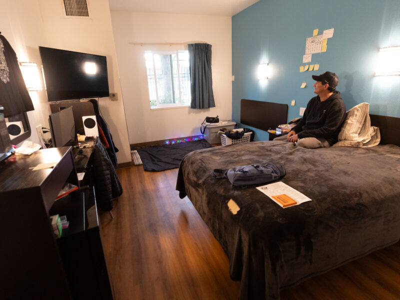 Wes sitting on his bed in a tidy motel room.